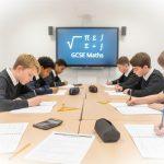 UK students studying for GCSE Maths exams with books, calculators, and equations on a classroom whiteboard representing the traditional learning route.